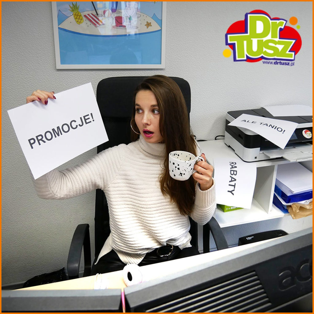 Kobieta siedząca na biurowym krześle ze zdziwioną miną trzyma kubek i kartkę z napisem PROMOCJE!